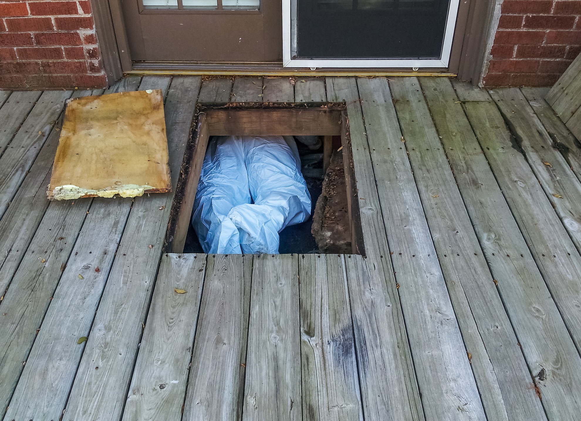 Workman with protective suit crawing under house from crawlspace underneath a wooden deck
